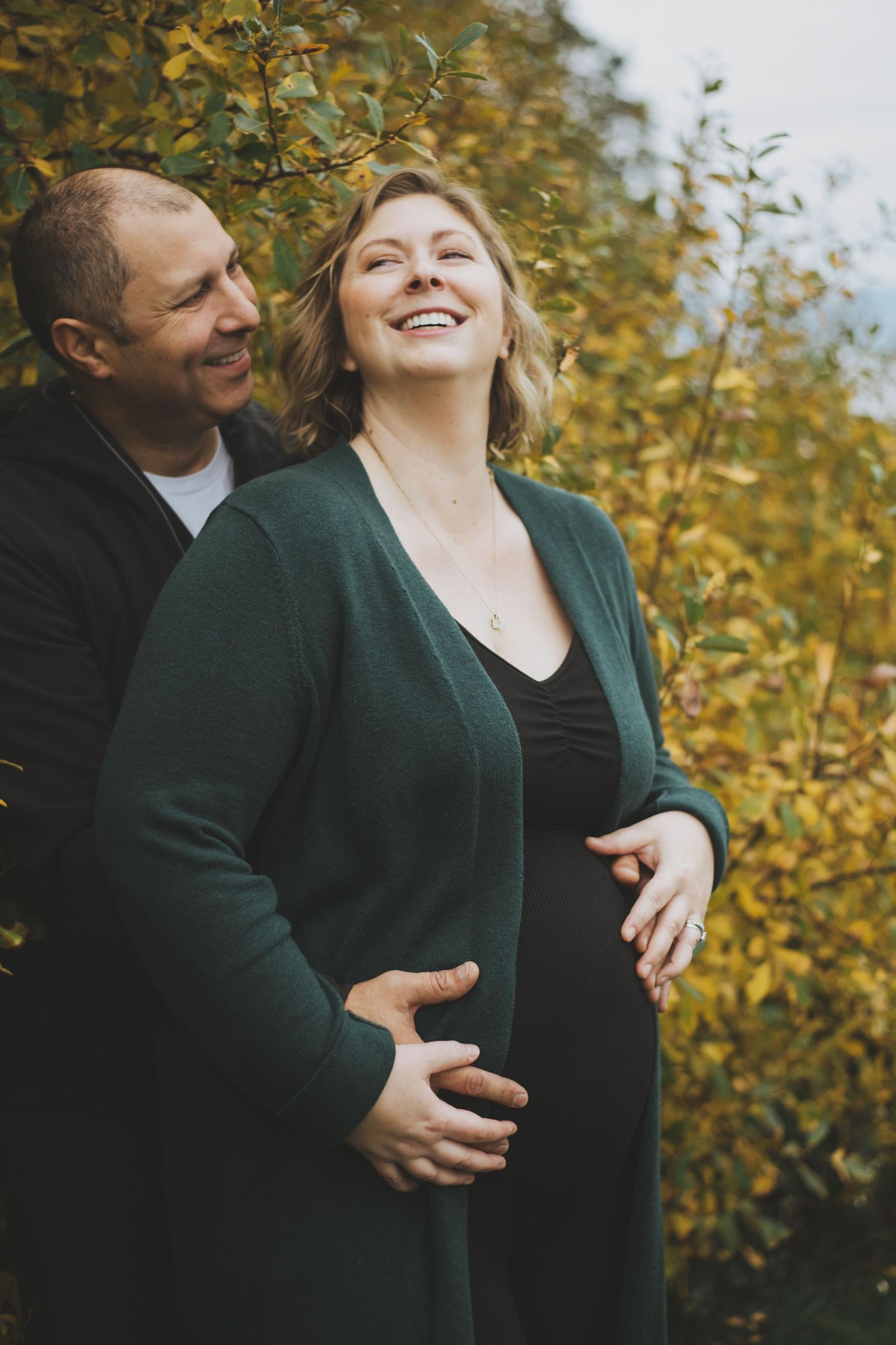 A couple shares a joyful moment together outdoors with autumn foliage in the background during a maternity photoshoot.