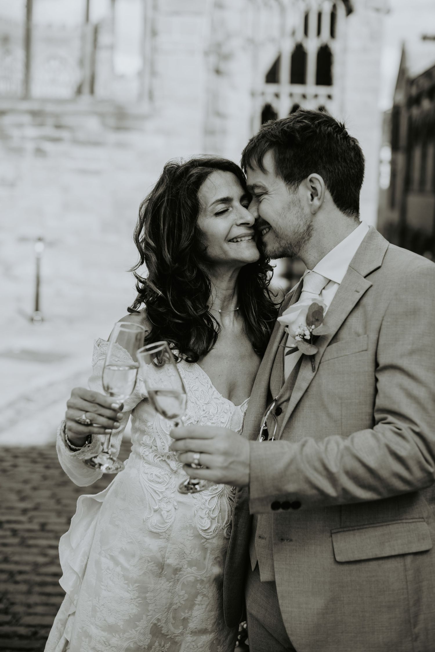 A tender black and white wedding moment shows newlyweds sharing champagne and embracing on a cobblestone street.