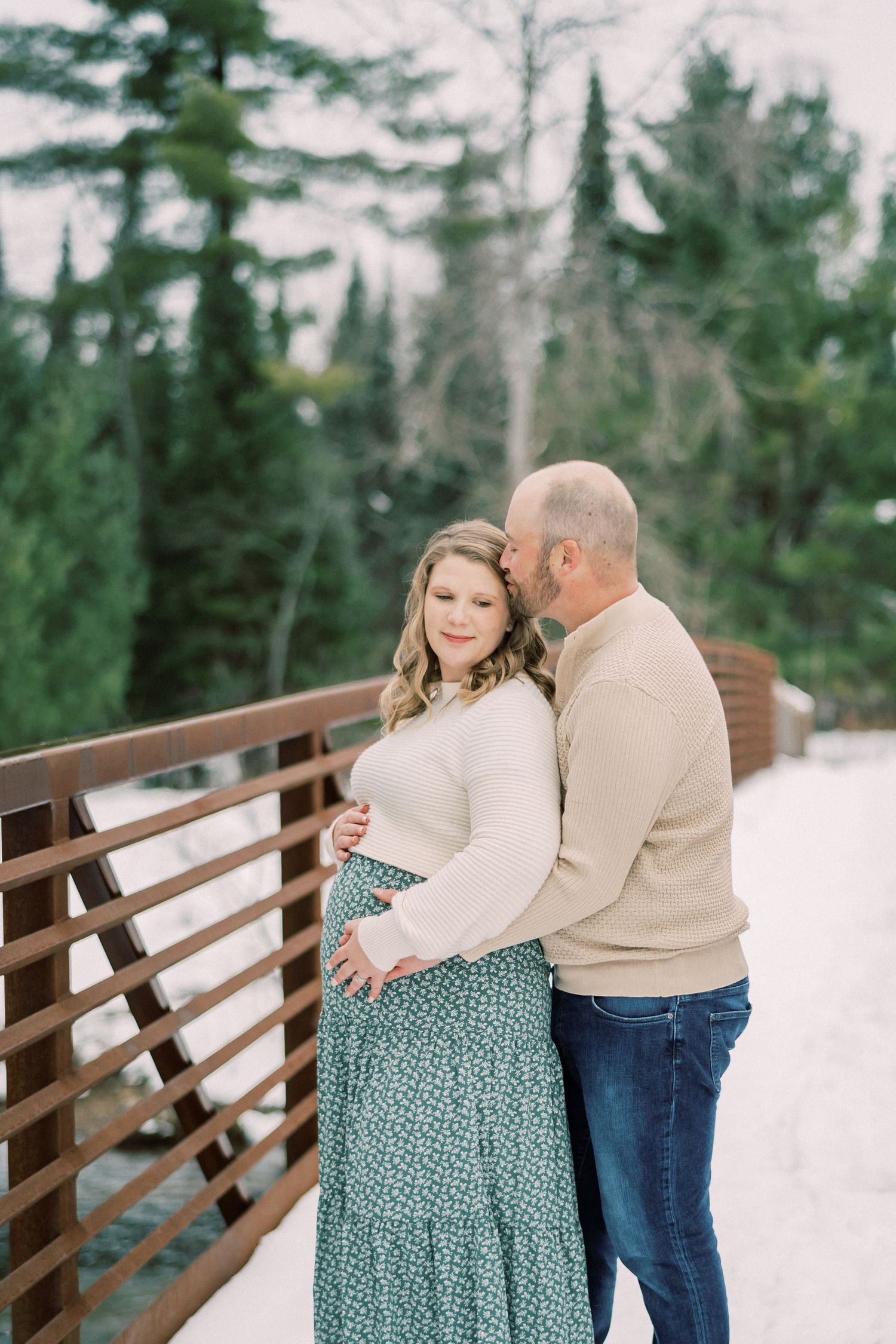 Intimate winter maternity photo on wooden bridge surrounded by snow-covered evergreens.