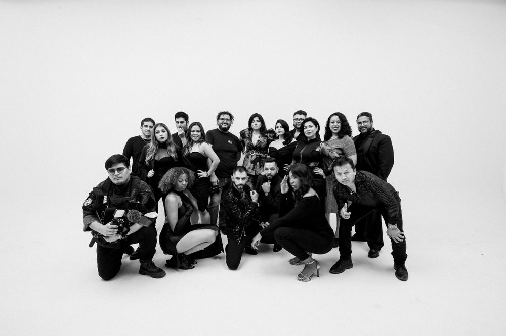 A large group poses together wearing all black outfits on a white background for a dramatic black and white photo.