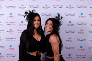 Two people wearing black outfits and feathered headpieces pose together at a branded step-and-repeat backdrop.