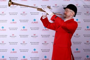 A musician in traditional red ceremonial uniform plays a trumpet against a branded backdrop with sponsor logos.