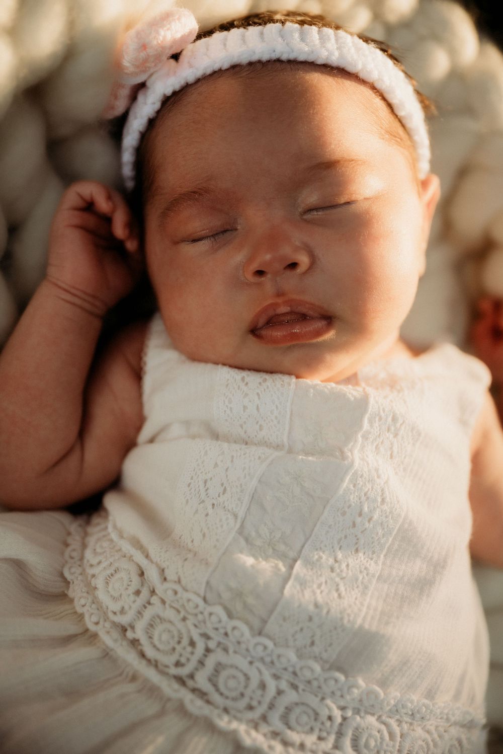 Baby wearing white headband and lace dress sleeping peacefully in soft lighting.