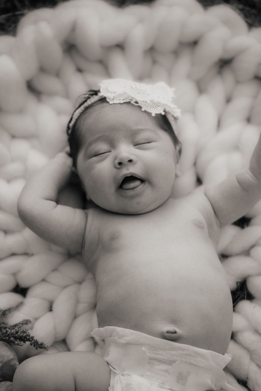 Baby with lace headband stretching on chunky knit blanket in black and white photo.