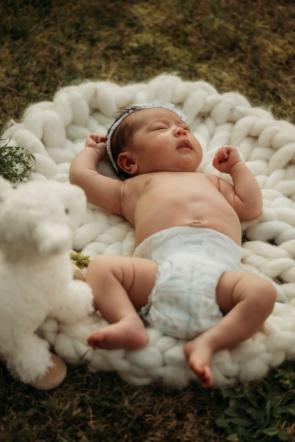 Infant wearing white headband stretches on chunky knit blanket outdoors.