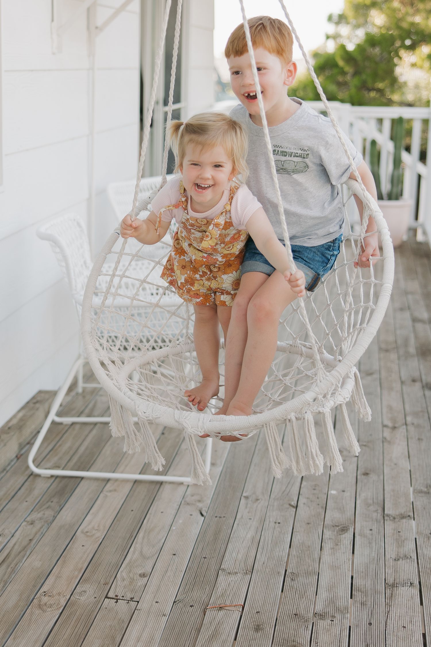 Two children playing and laughing in a white macrame hanging chair on a porch.