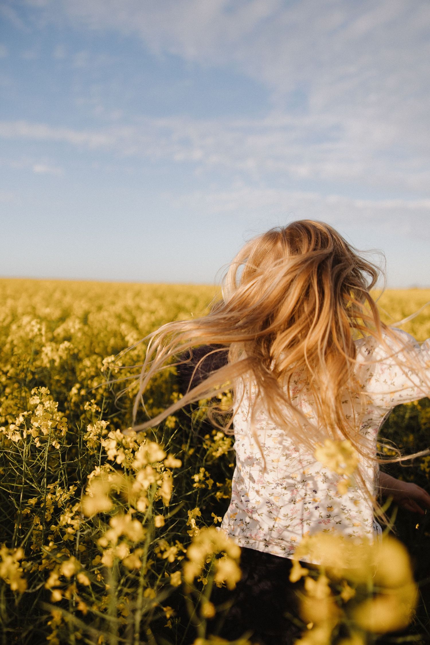 Woman with flowing hair in floral dress spinning in yellow canola field.