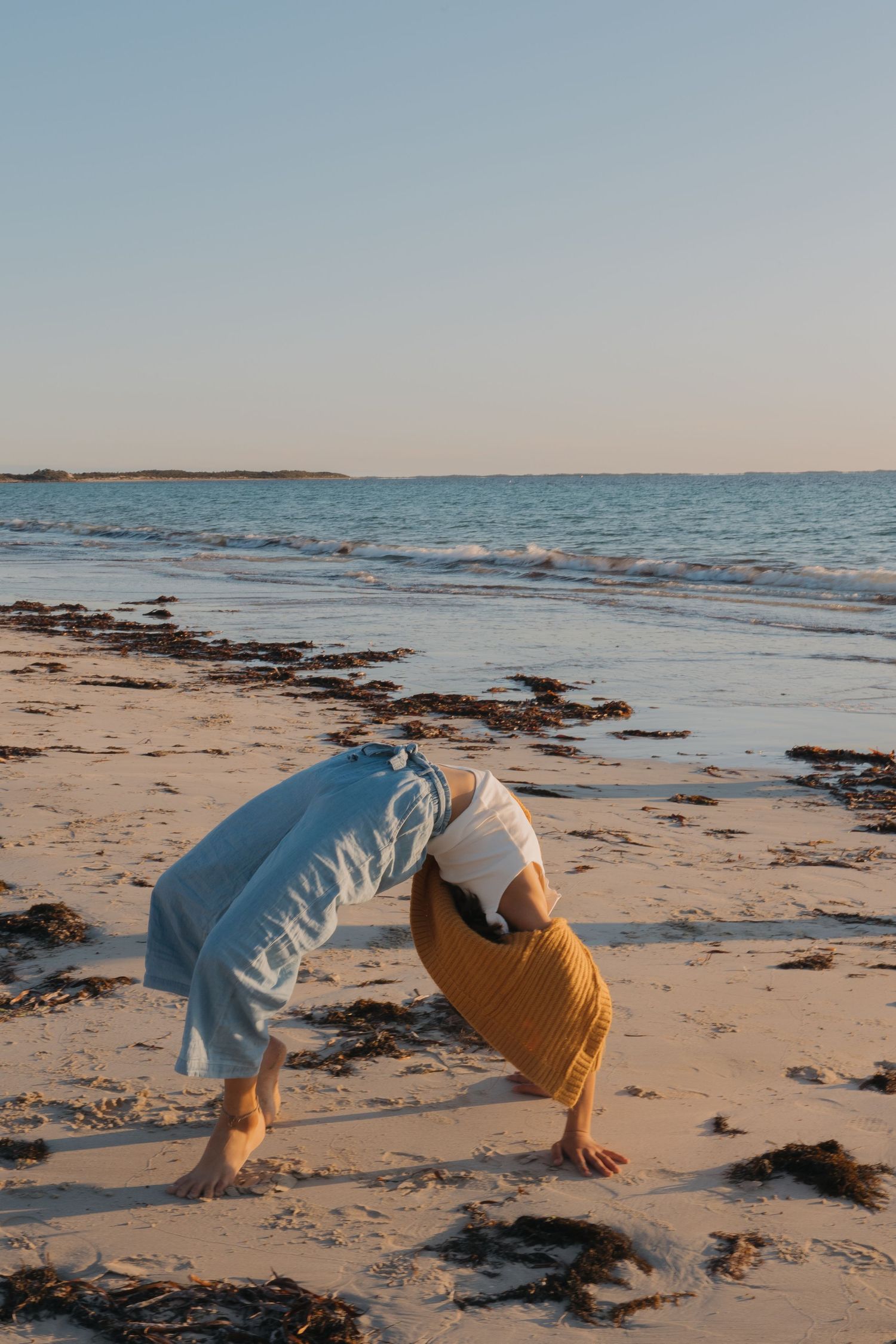 Two figures in casual clothing playfully bend over on a beach at sunset.