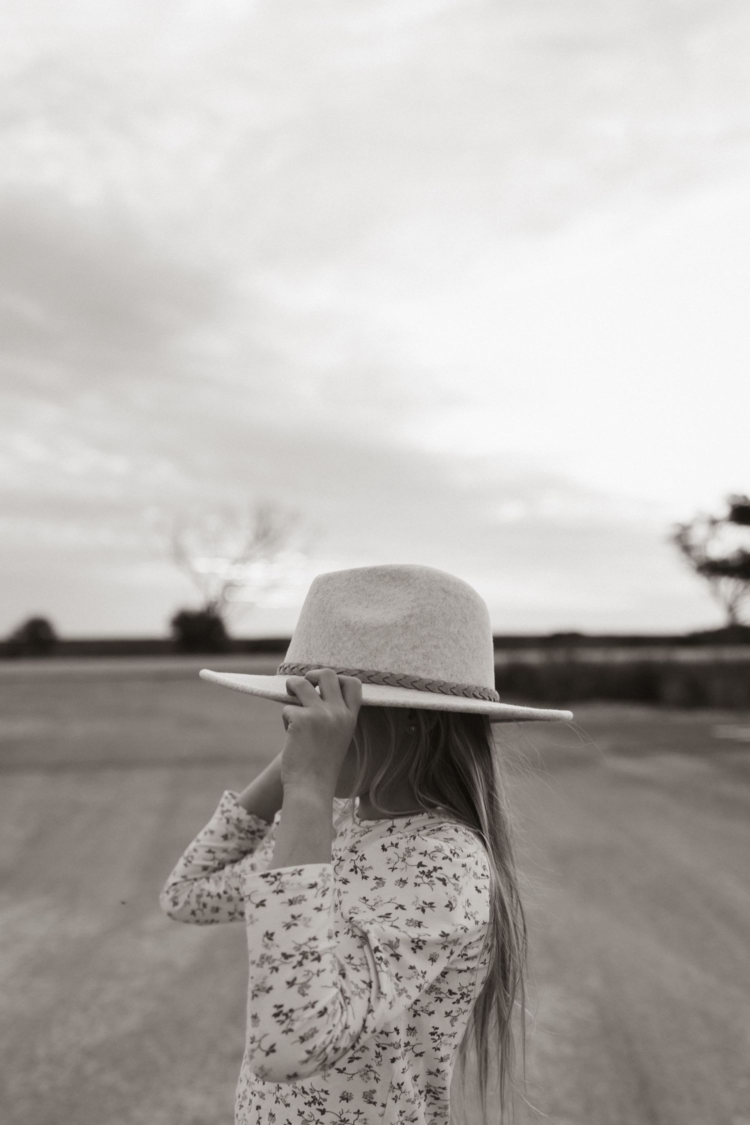 Person in floral dress and hat standing on rural road in black and white.