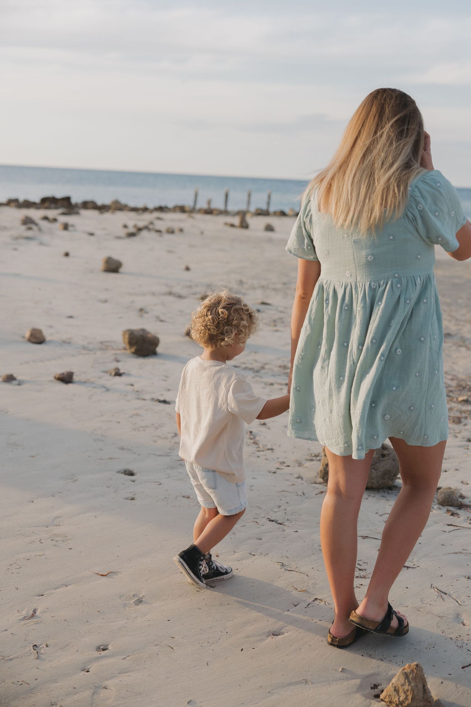 A parent and child walk hand in hand along a sandy beach with rocks and posts in the distance.