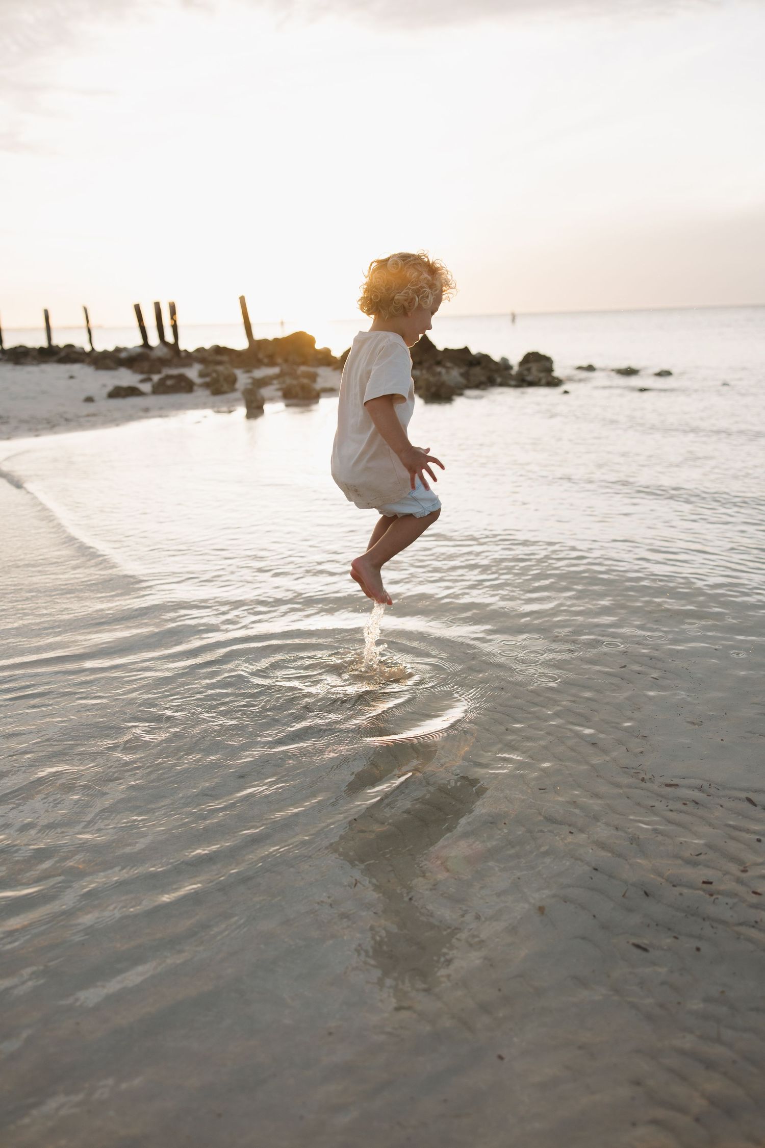 A child jumps playfully in shallow beach water at sunset creating splashes.