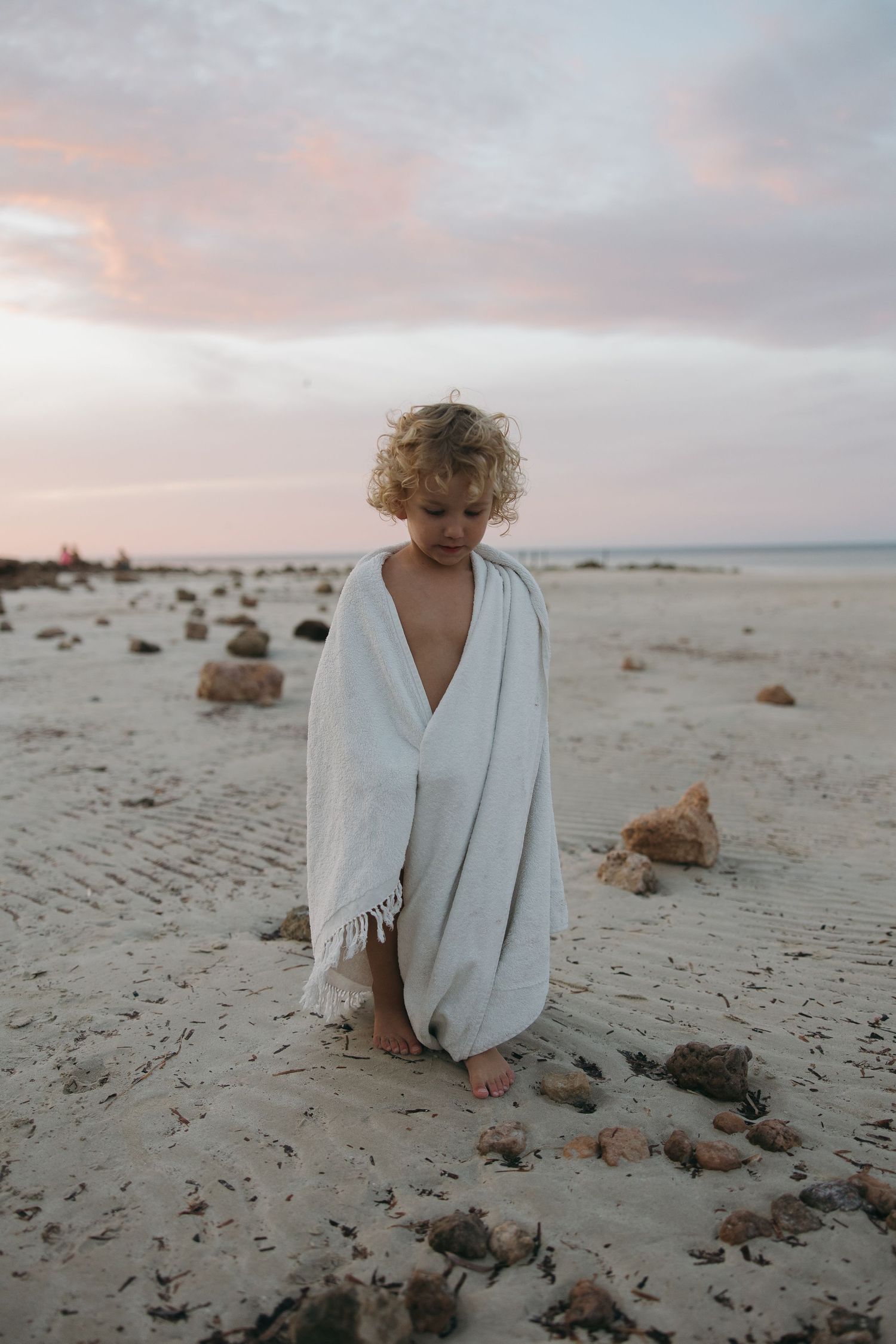 A figure wearing a white flowing dress stands on a rocky beach at sunset.