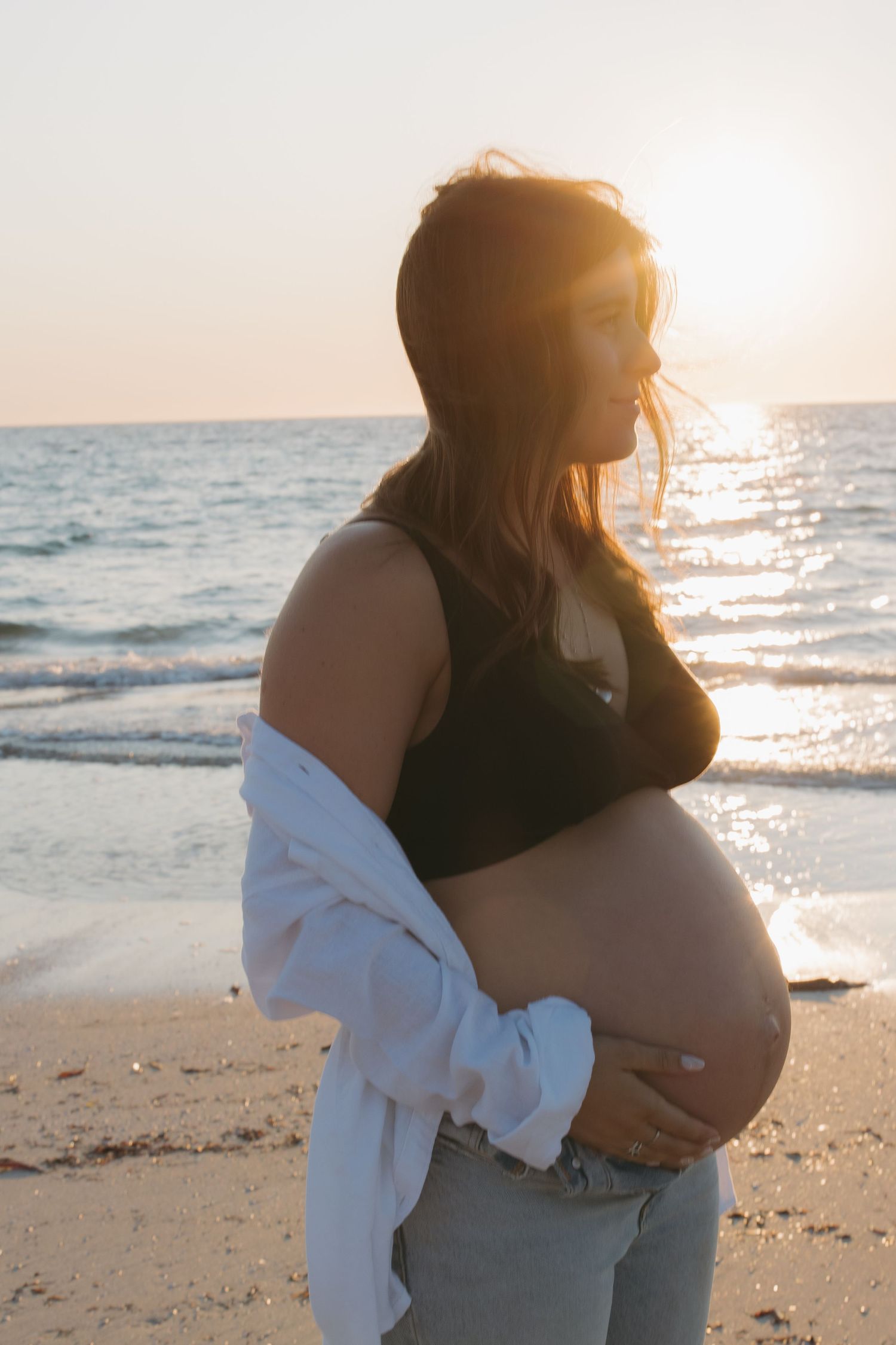 A pregnant woman in casual attire stands on the beach during golden hour, cradling her baby bump.
