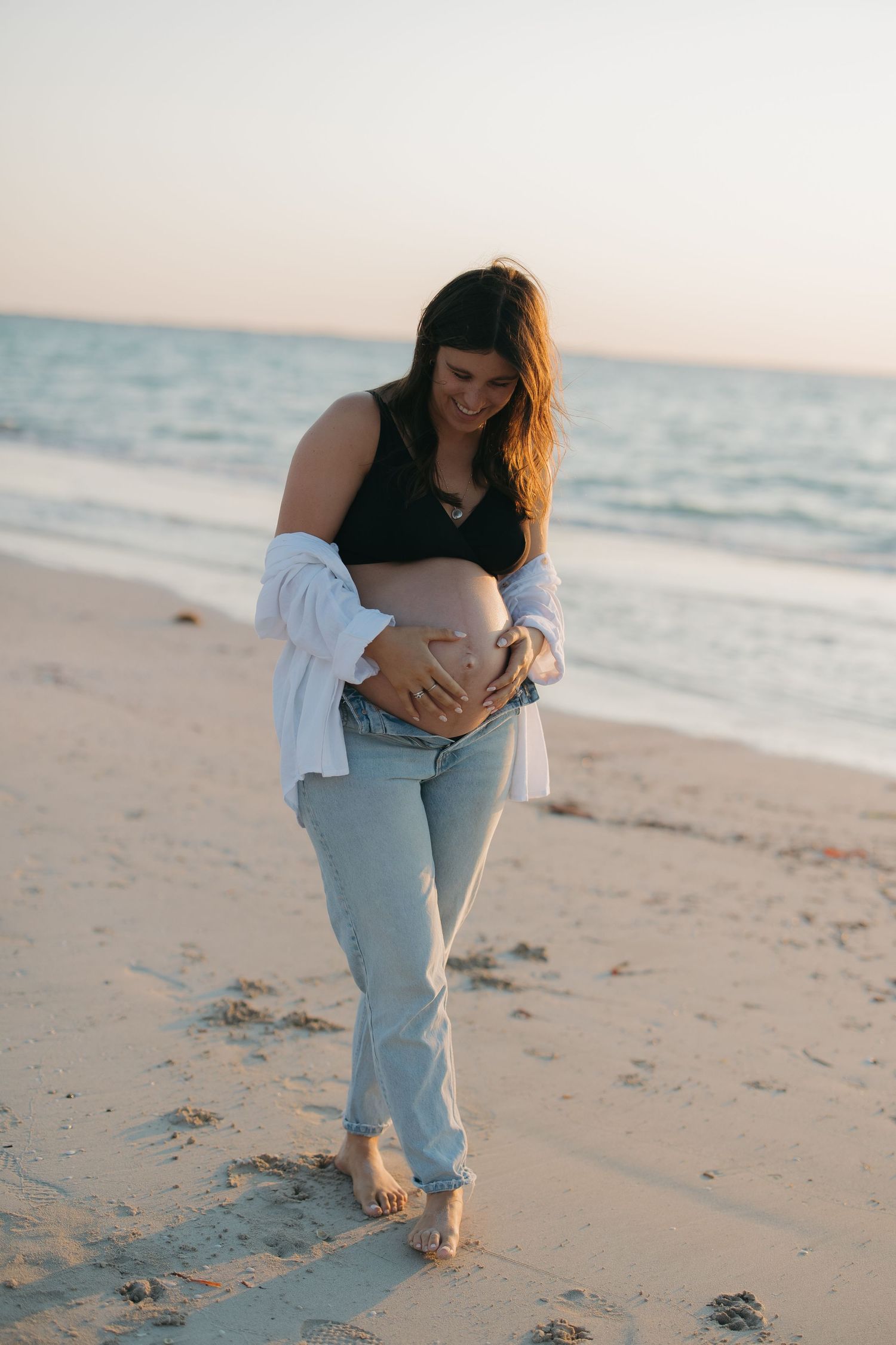 A pregnant woman in light jeans and white cardigan walks barefoot along a serene beach at sunset.