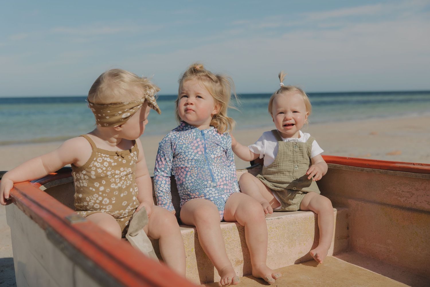 Three toddlers sitting together in a wooden boat on a sunny beach.
