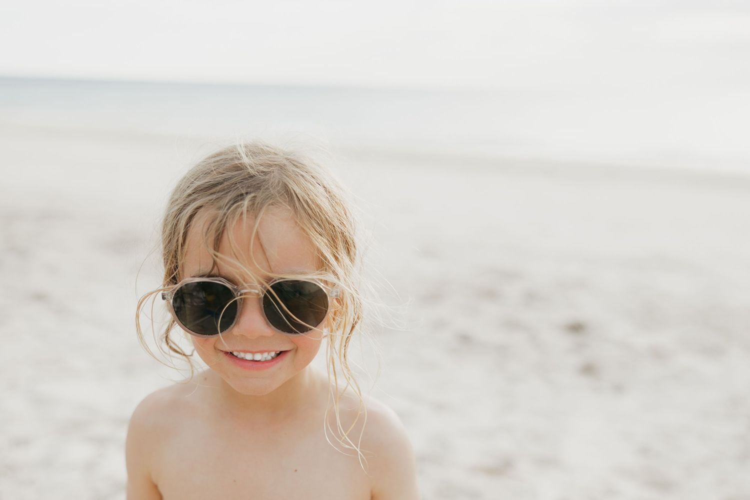 A child wearing aviator sunglasses smiles brightly on a sunny beach.