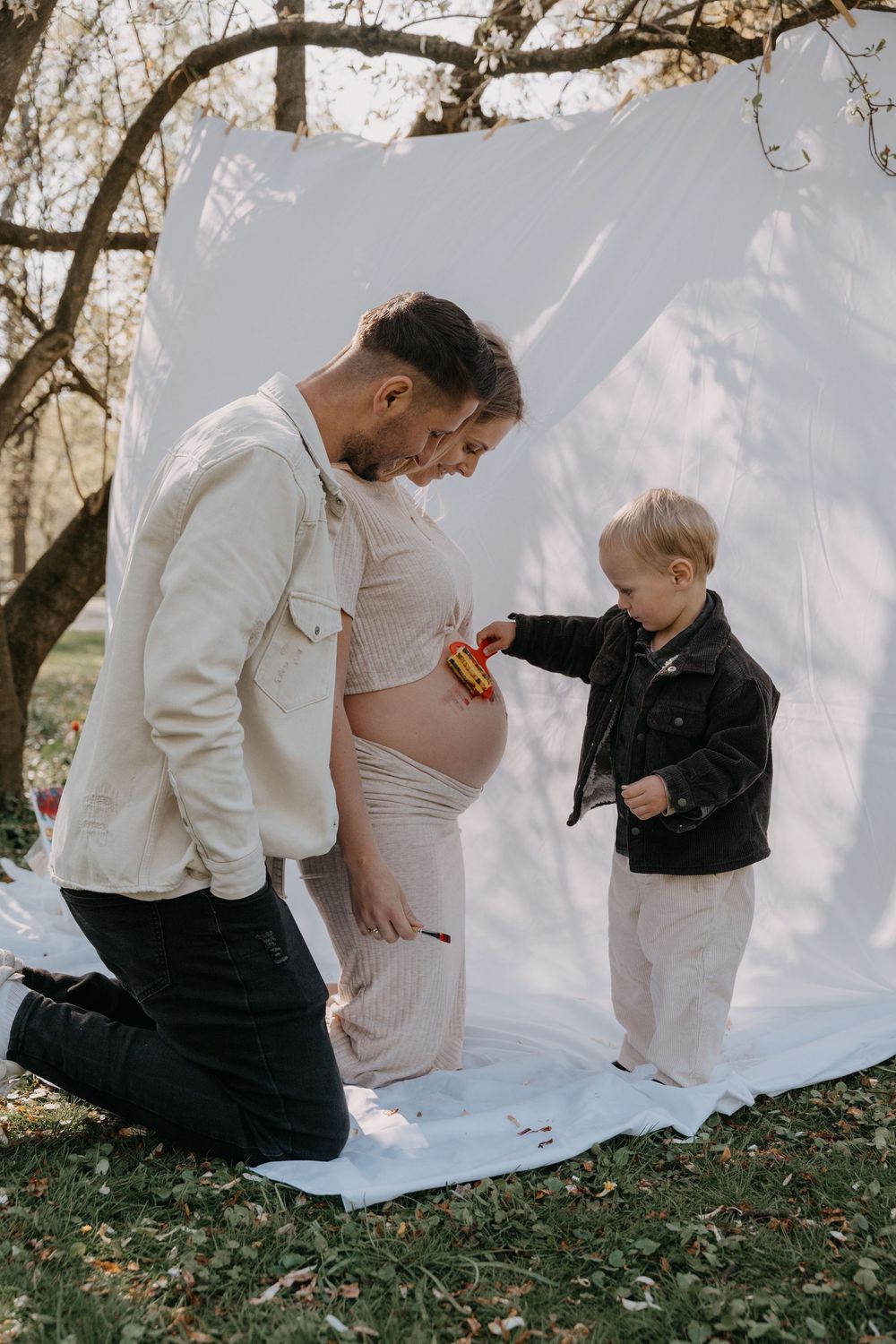 A tender family moment outdoors with a pregnant woman in white as a young child touches her baby bump against a light backdrop.