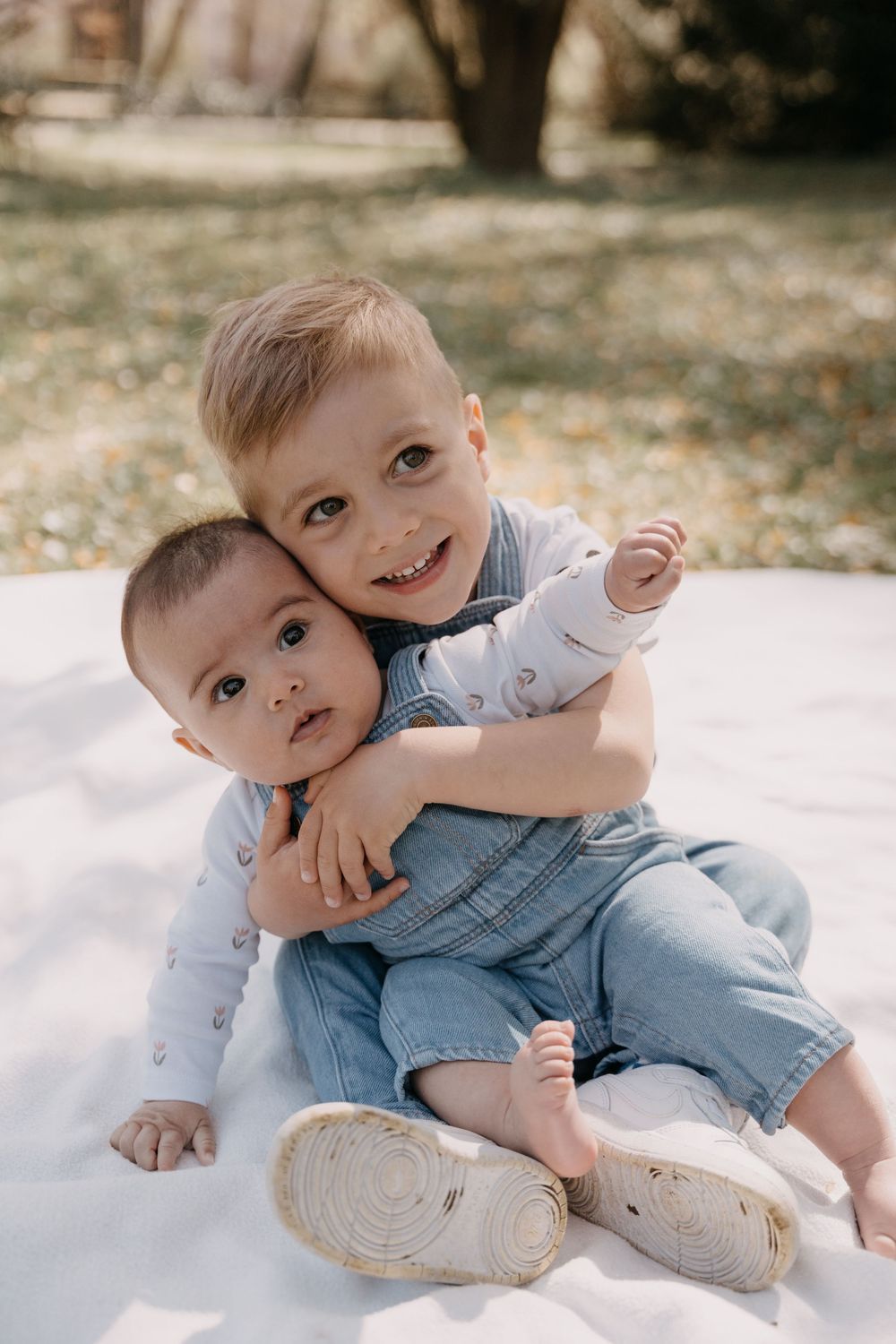 A happy toddler in denim overalls lays on a white blanket in an outdoor setting showing playful expressions.