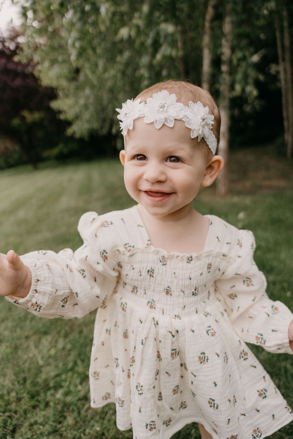 Sweet baby wearing white floral dress and delicate lace headband smiles brightly in an outdoor garden setting.