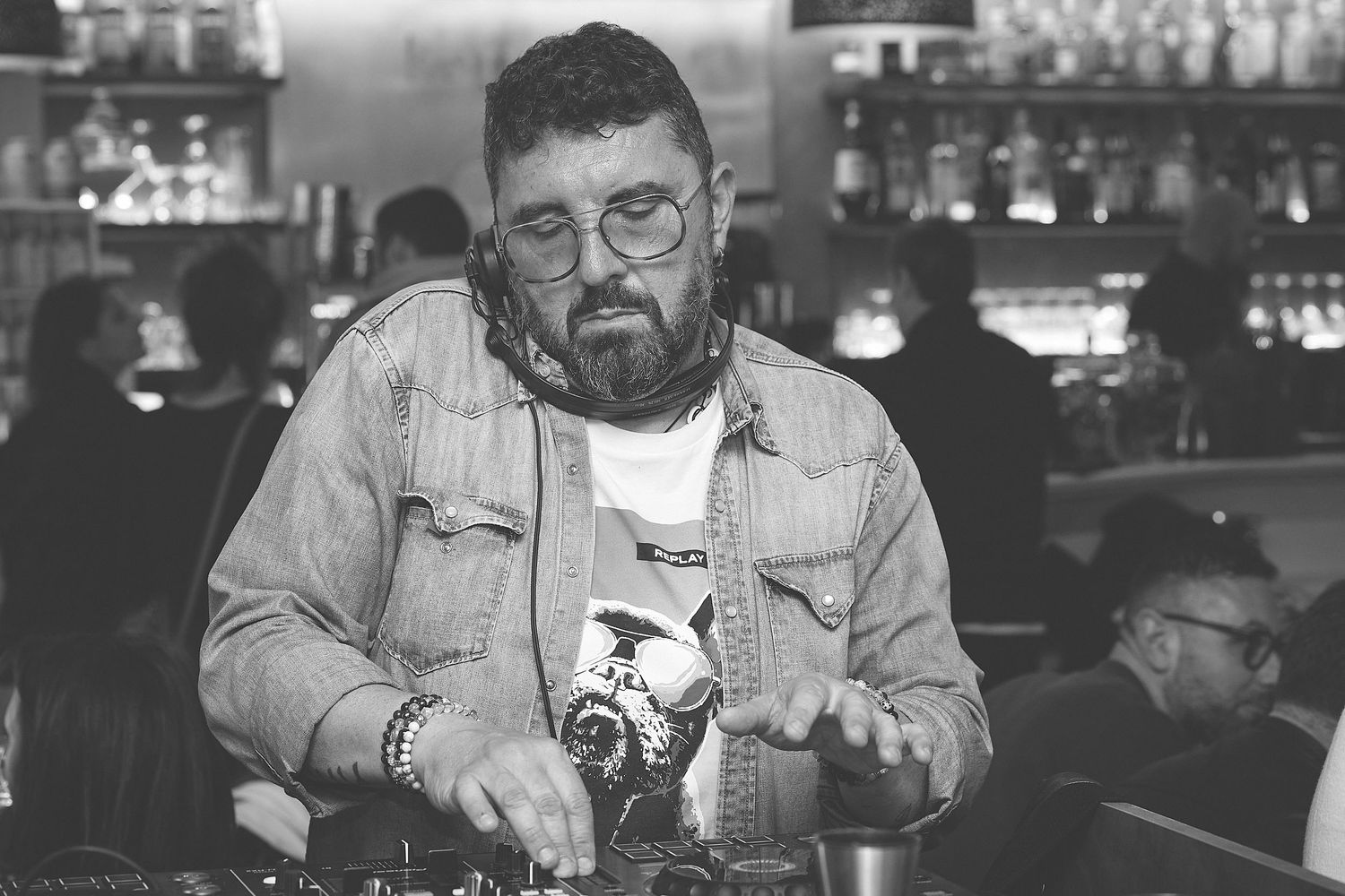 Bartender wearing denim and glasses mixing drinks at a bar counter in black and white.