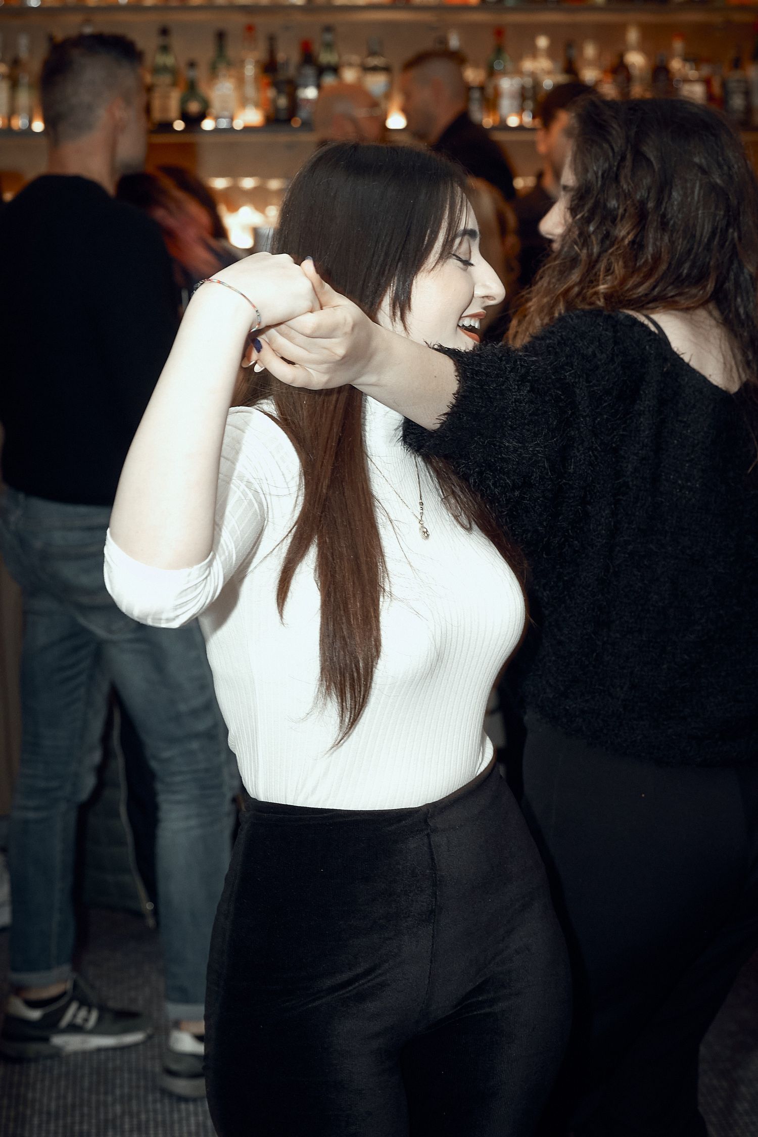 Woman in white sweater and black pants dancing at a crowded bar.