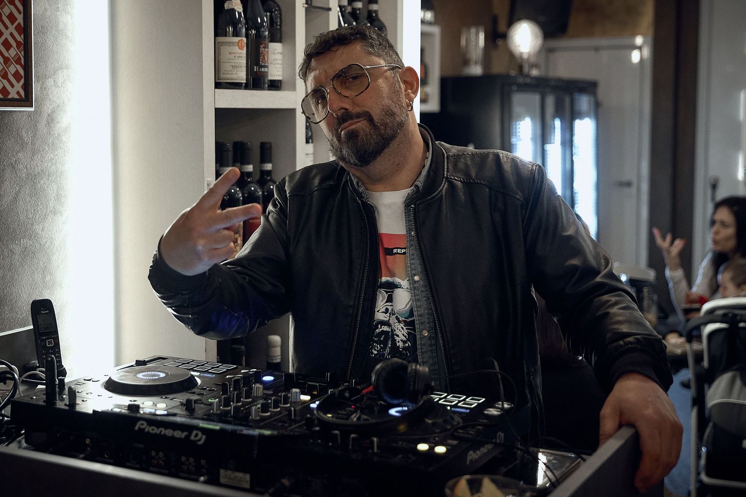 DJ in leather jacket mixing music at turntable setup in dimly lit venue.