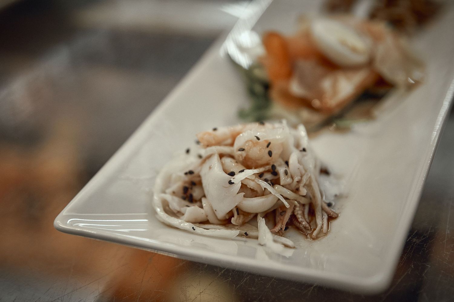 Close-up of noodle dish garnished with shaved toppings on white rectangular plate.