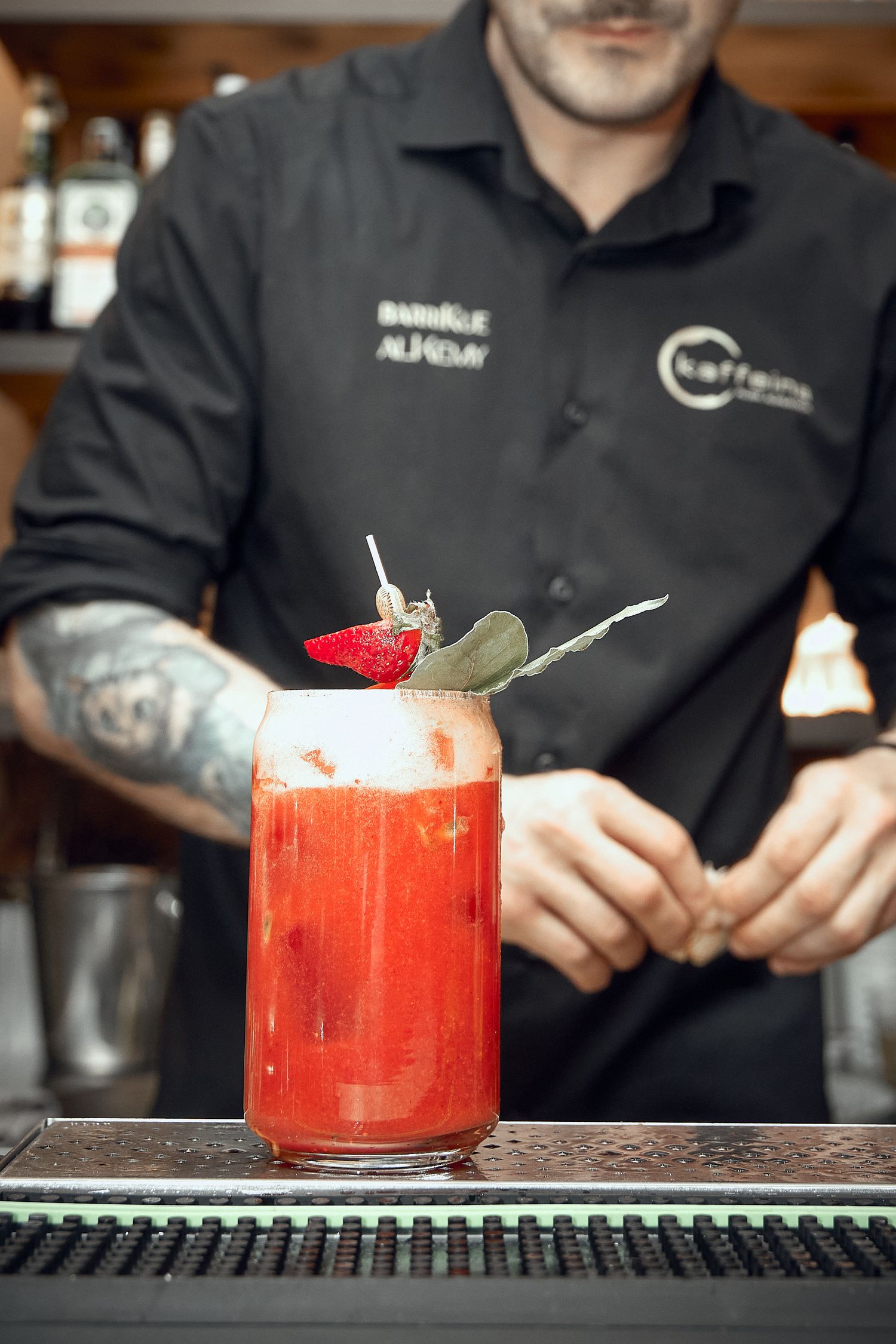 Bright red cocktail garnished with strawberry being prepared at bar counter.