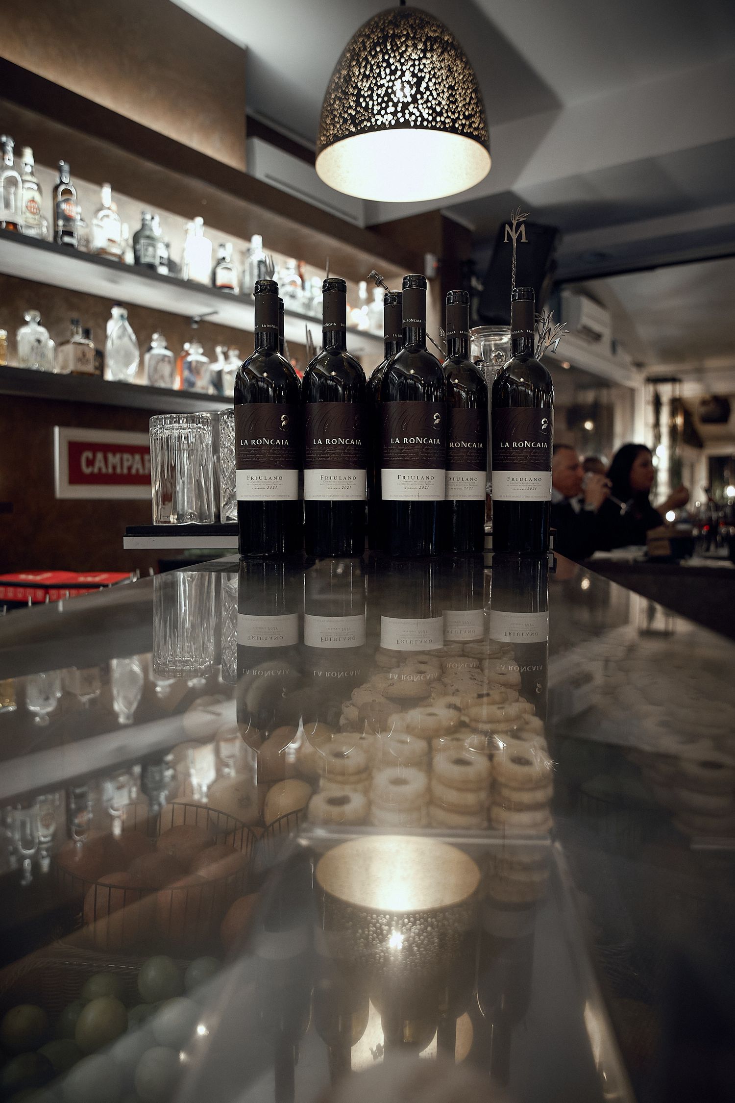 Wine bottles lined up on reflective bar counter under decorative pendant light.