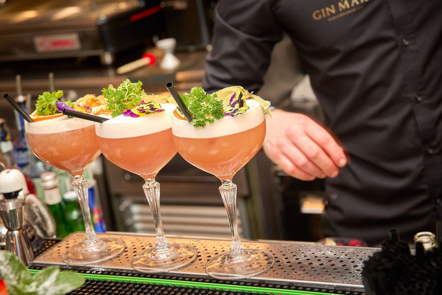 Three pink cocktails garnished with fresh herbs lined up on a bar counter.