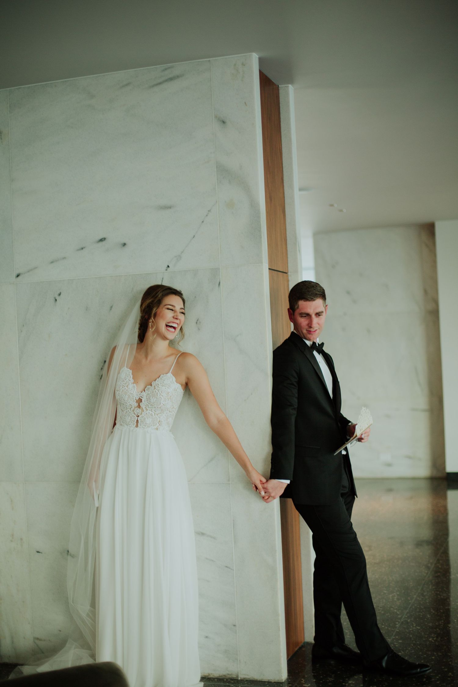 A joyful couple shares a playful moment while holding hands against a marble wall.