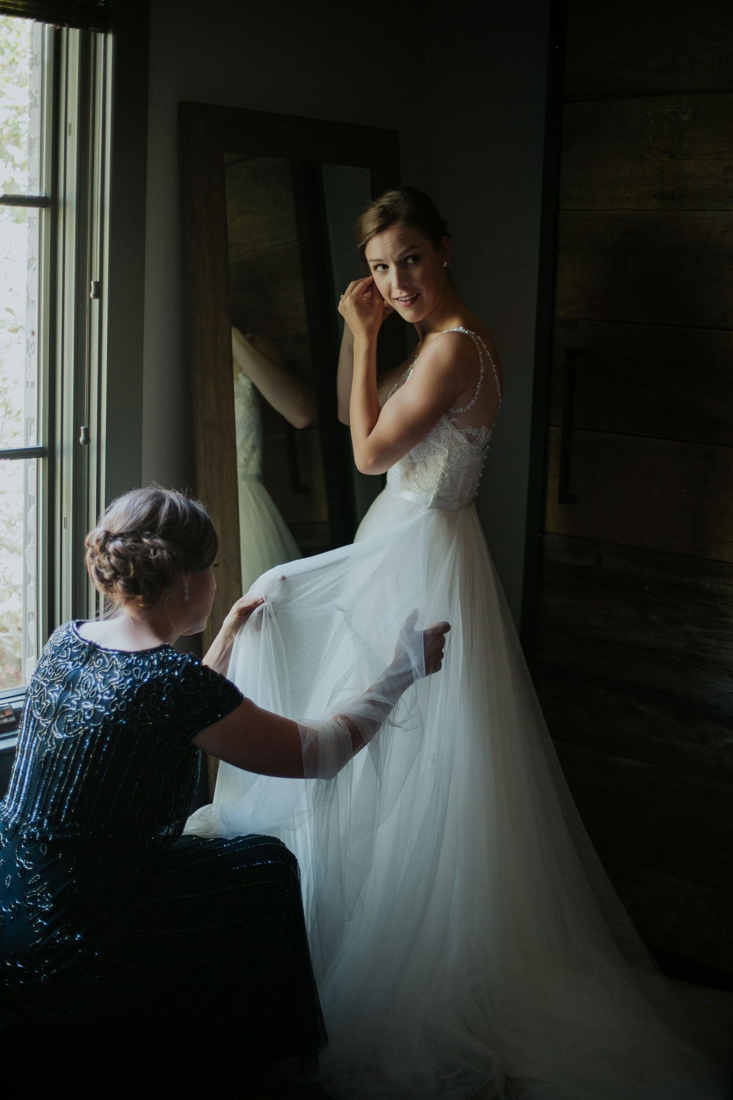 A touching moment as someone helps adjust a white wedding dress by a window in soft natural light.