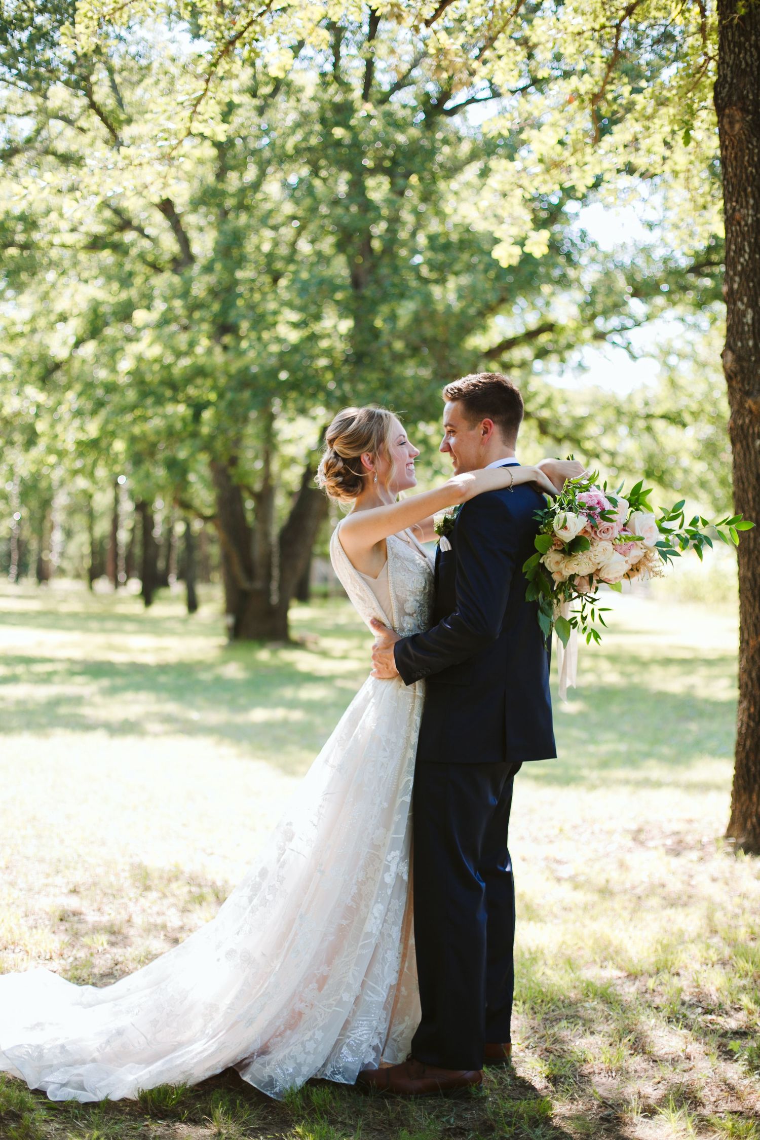 A romantic wedding couple embraces outdoors among sunlit trees with the bride holding a floral bouquet.
