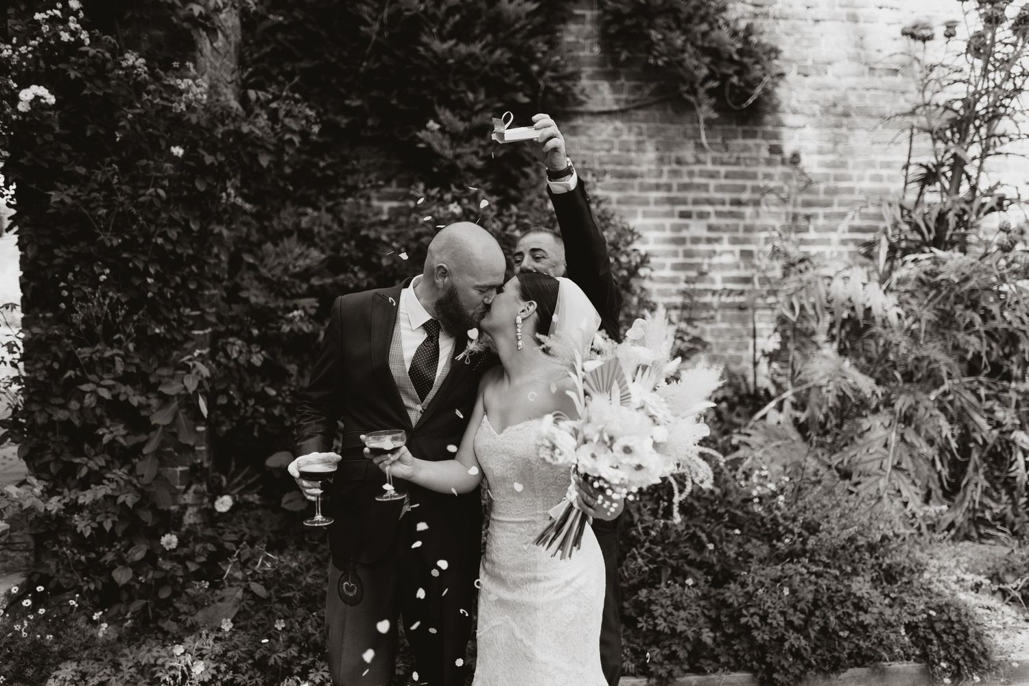 A bride in white walks through a scenic garden pathway bordered by lush greenery and a brick wall.