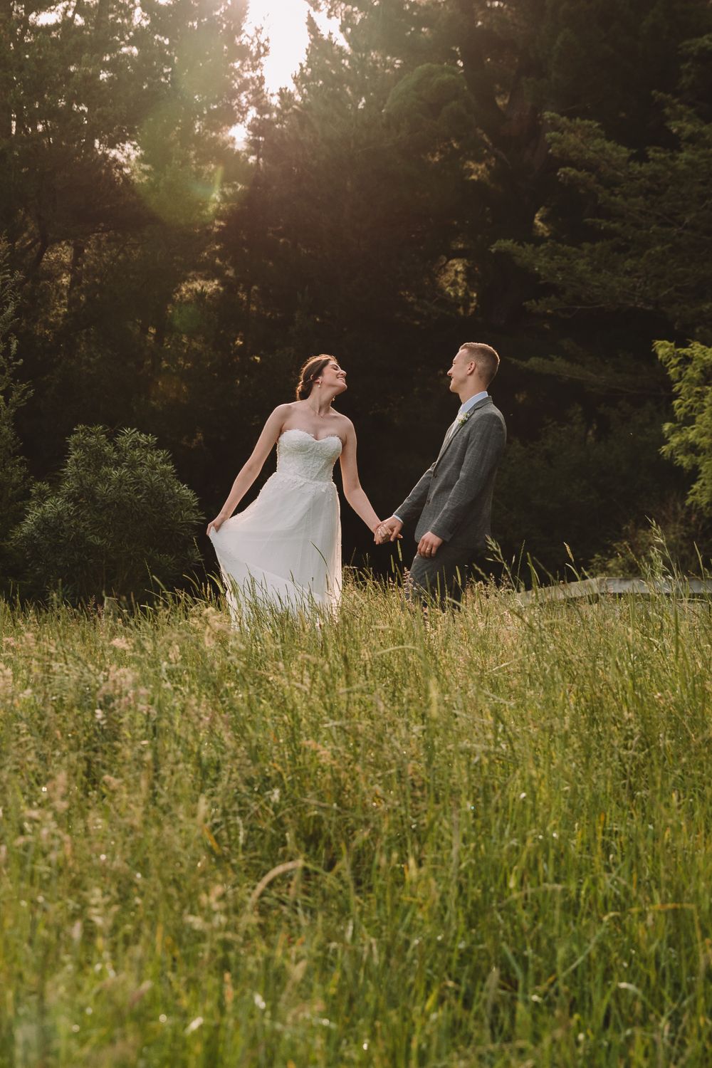 Sunset wedding photo of couple standing in tall golden grass with forest behind.