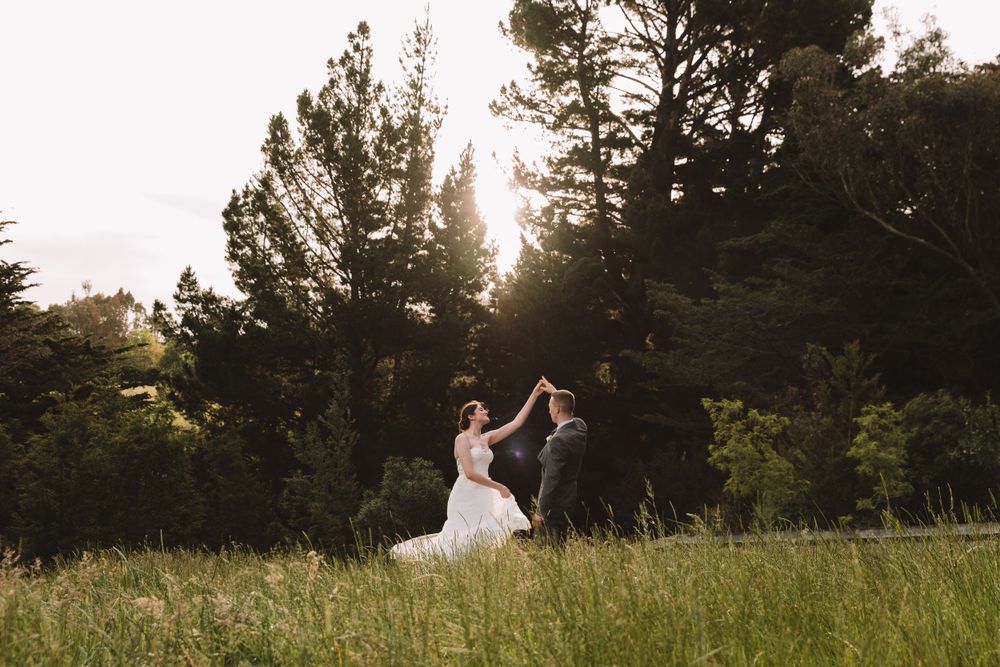 Couple dancing in a sunlit meadow with evergreen trees in the background.