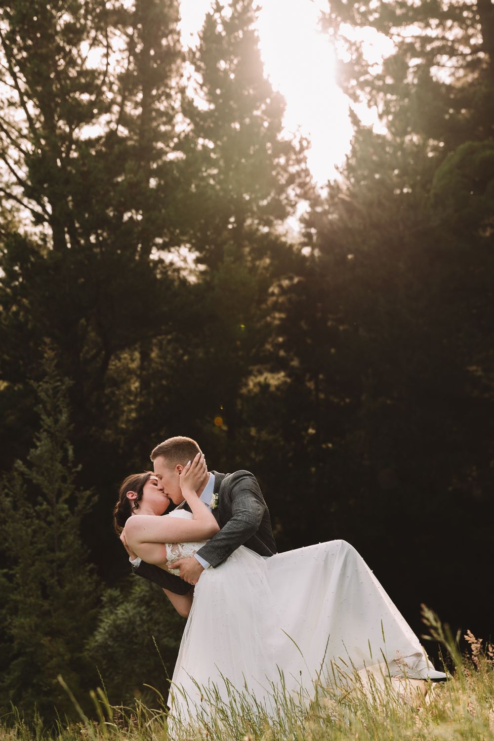 A romantic wedding couple share a passionate kiss in a forest at sunset with dreamy backlighting.
