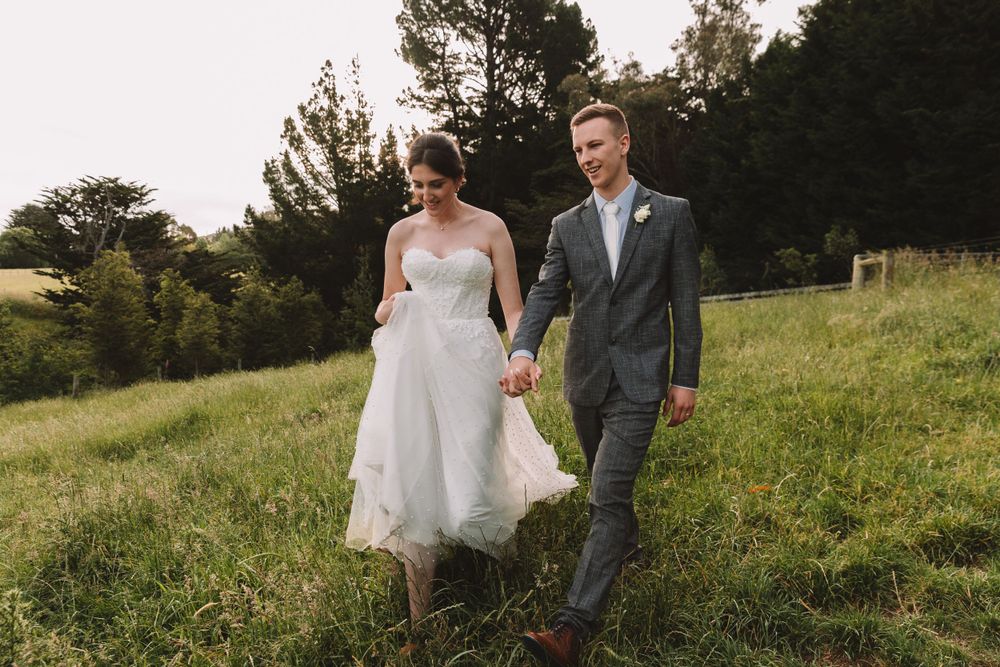 A couple in wedding attire walking through a lush green meadow during golden hour.