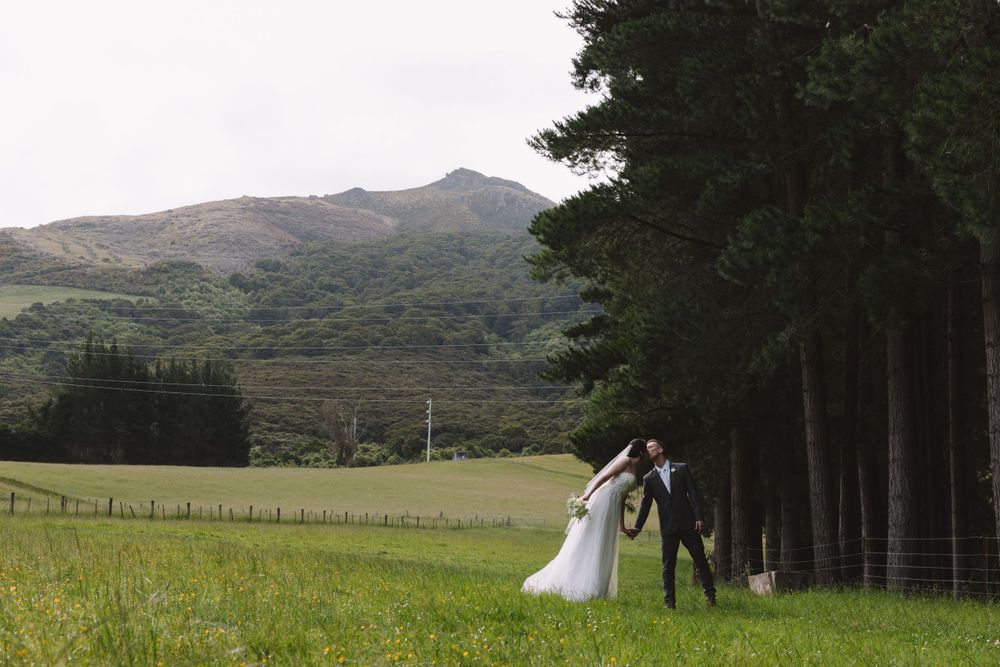 A couple in wedding attire shares a romantic moment near a line of evergreen trees with mountains behind them.