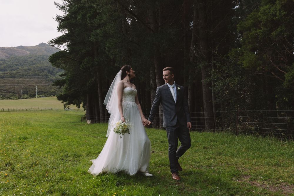 A couple walks hand in hand through a lush green field with mountain scenery in the background.