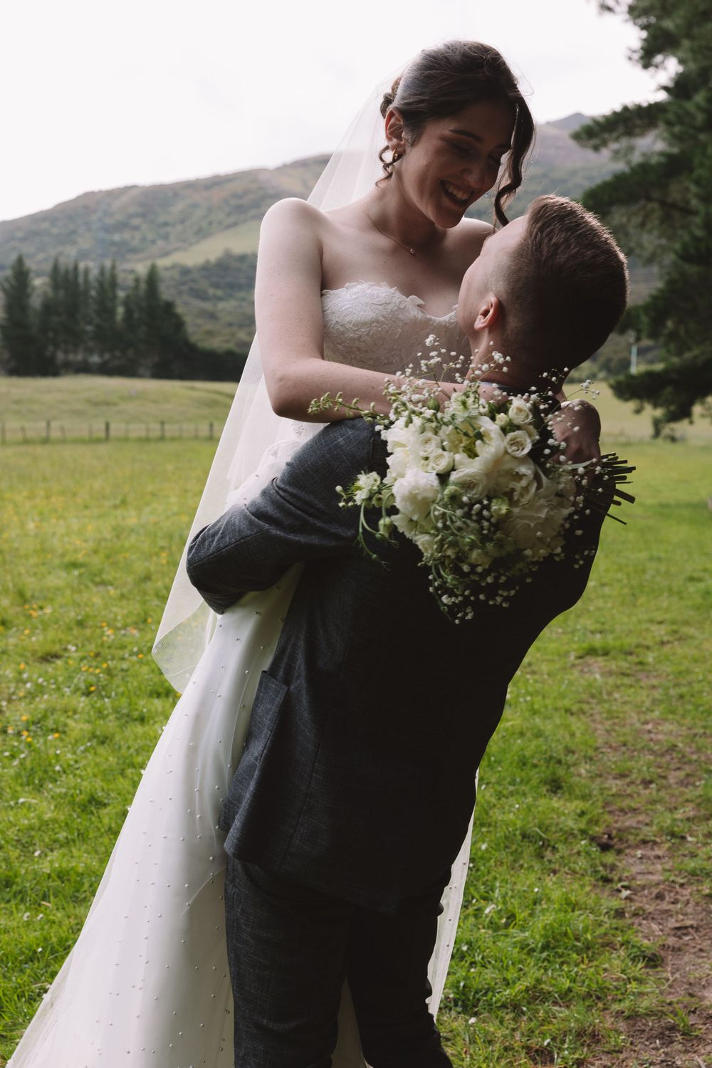 A romantic embrace between newlyweds with a white floral bouquet in a mountain setting.