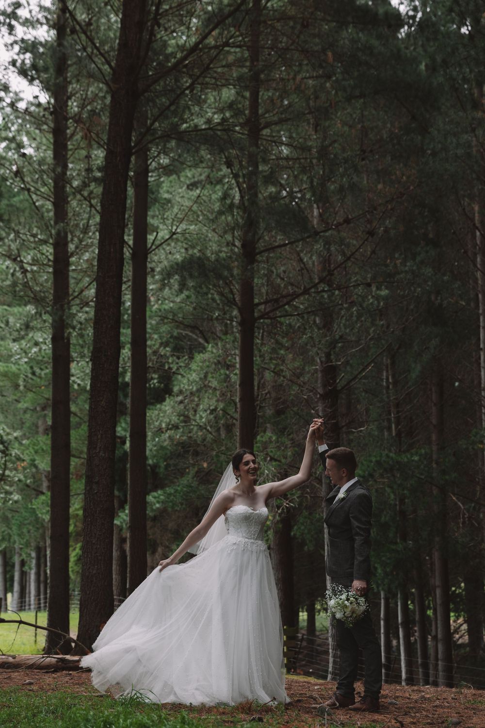 A romantic wedding moment captured between pine trees as a couple dances in an enchanted forest setting.