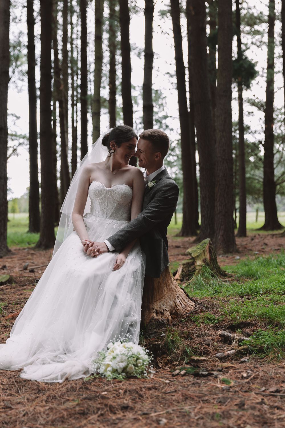 A tender wedding portrait taken in a pine forest with the couple sitting on a tree stump.