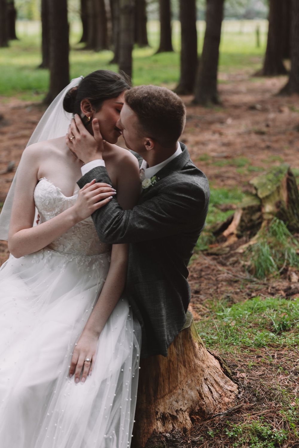 A passionate wedding kiss captured in a forest setting with tall pine trees in the background.
