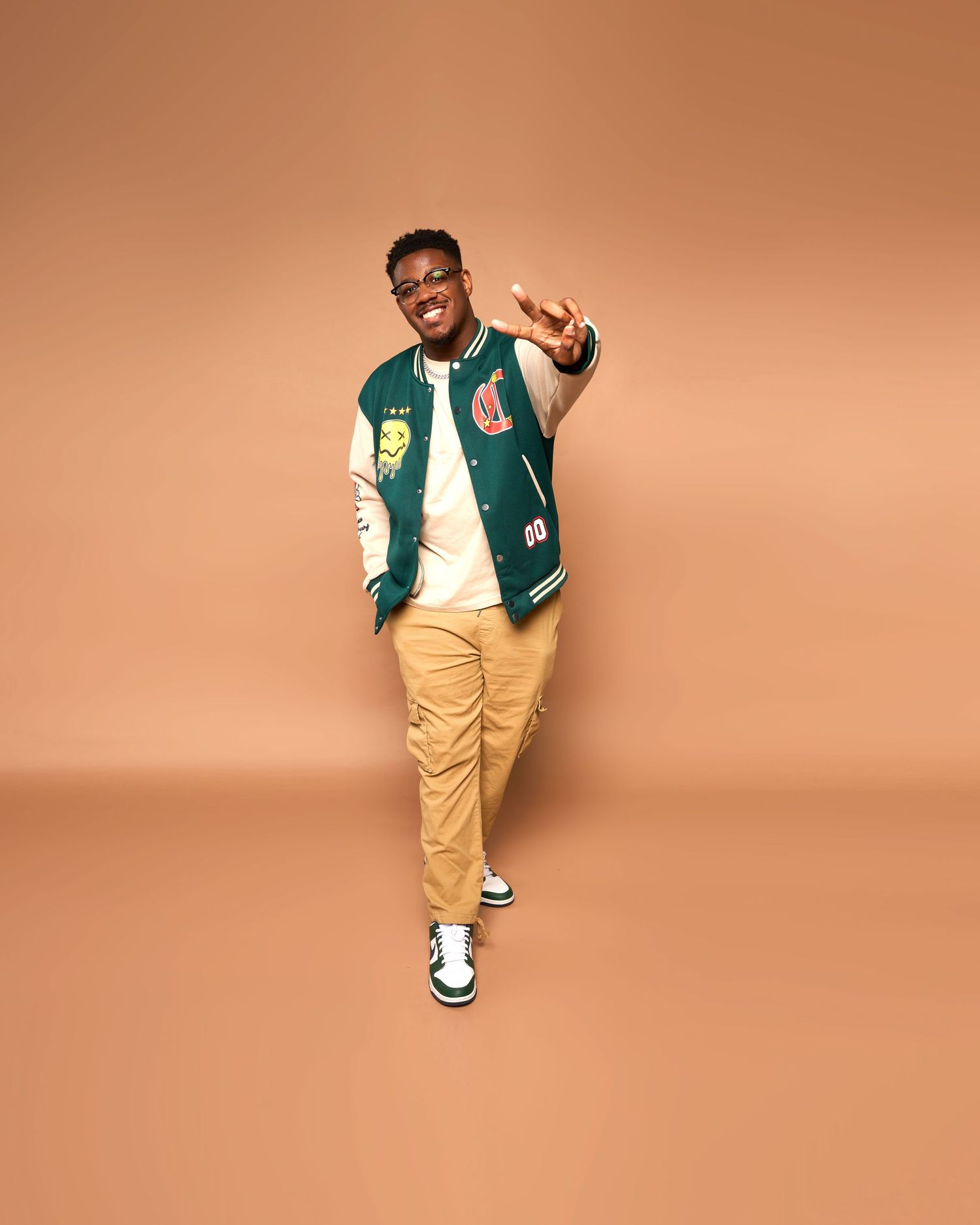 Studio portrait of person wearing varsity jacket and khakis posing against orange backdrop.