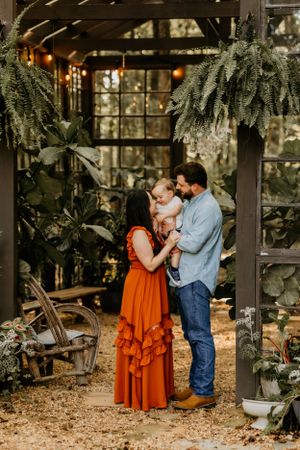 Family shares an intimate moment in a greenhouse filled with hanging plants and twinkling lights.