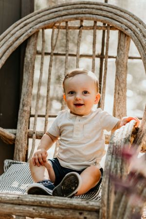 Happy toddler sits on rustic wooden chair in warm sunlit setting.