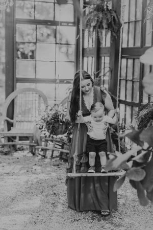 Black and white photo of parent and child playing on vintage wooden swing in a garden setting.