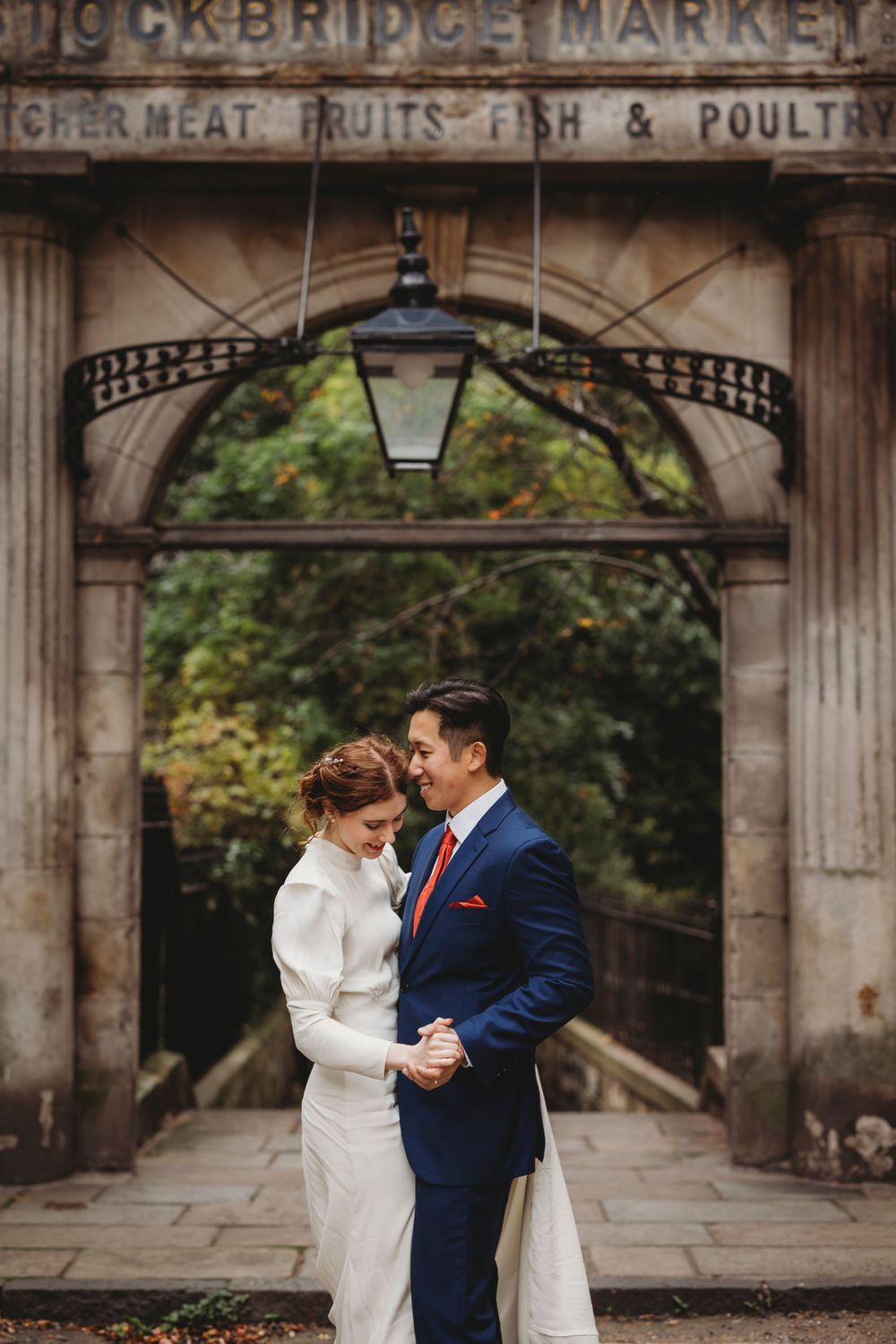 A romantic wedding couple embraces under the historic entrance archway of Stockbridge Market in Edinburgh.