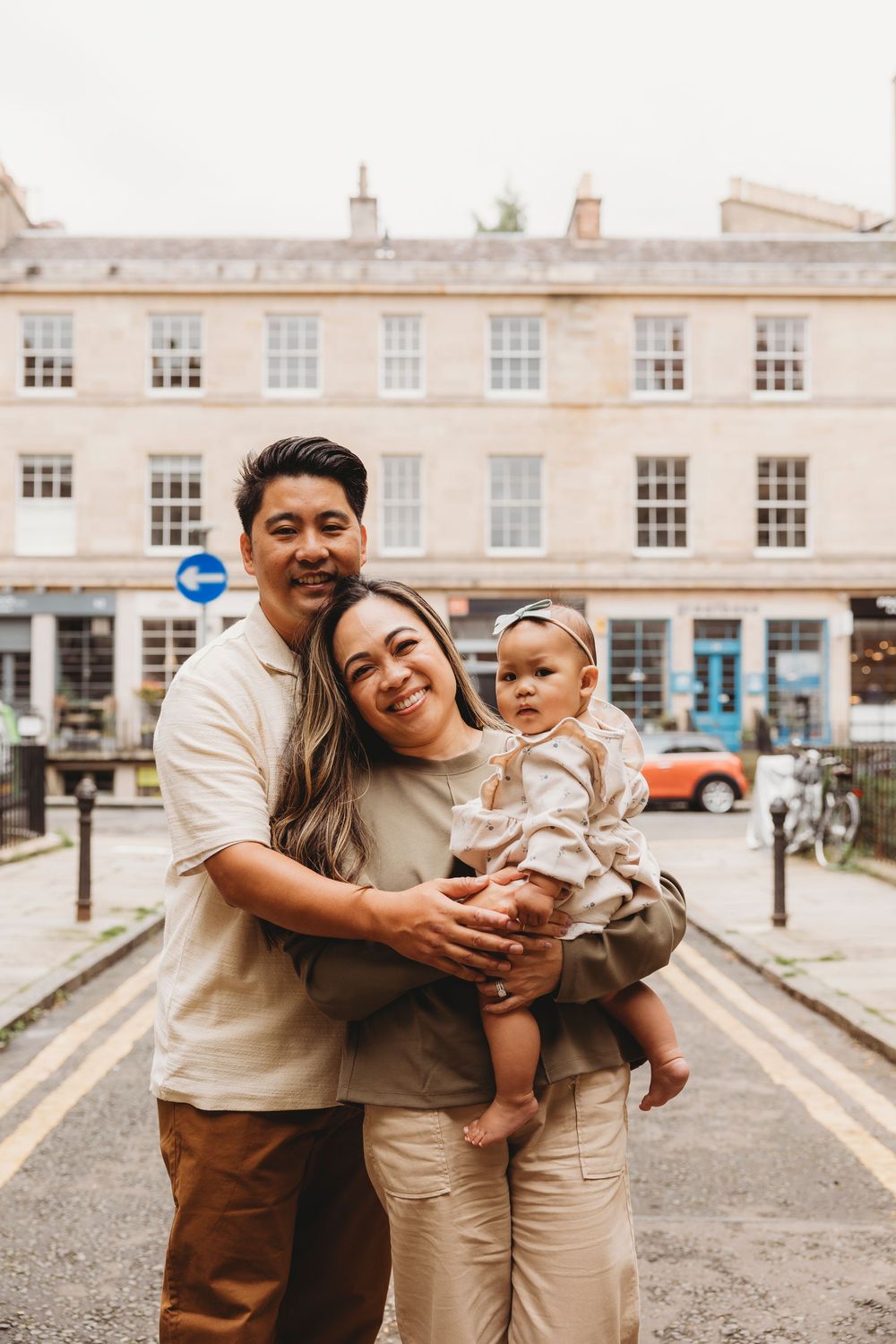A happy family stands together on a street in front of a historic Georgian building wearing neutral toned outfits.
